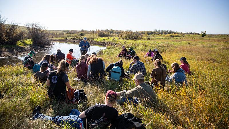 Enviro students sit in the grass by the river working on a lab